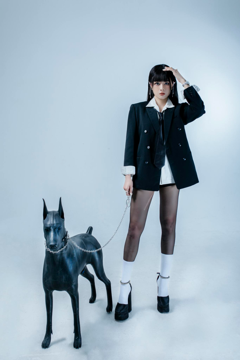 Asian woman in stylish black attire poses with a sleek dog statue in a studio.